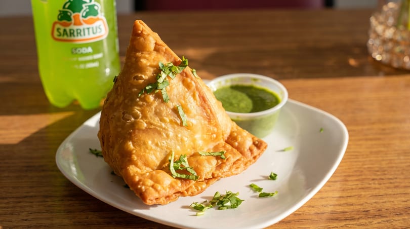Golden-brown samosa with cilantro on a white plate, green chutney, and a Sarritus soda.