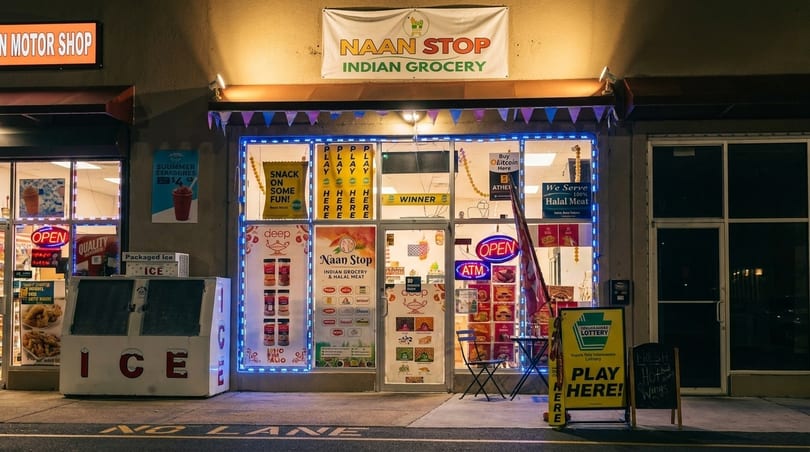 The brightly lit storefront of Naan Stop Indian Grocery, featuring various signs and products visible through the windows.